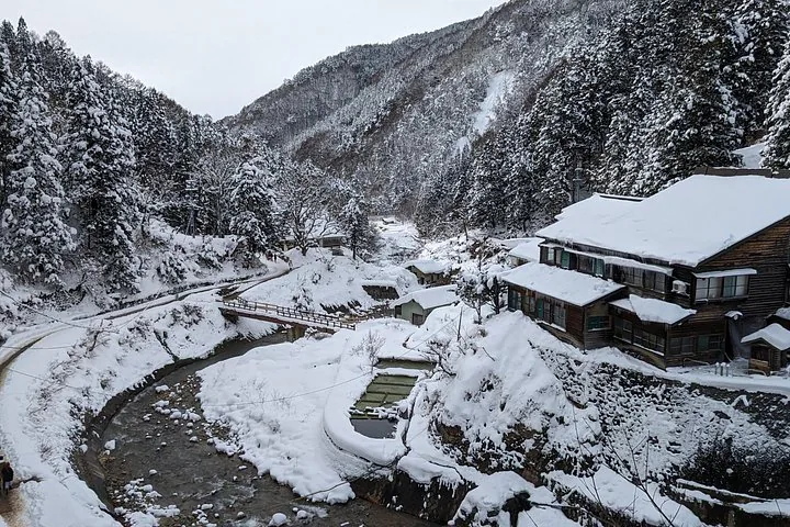 Eine verschneite Winterlandschaft mit einem Fluss, einer Brücke und traditionellen Holzgebäuden.
